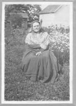SA0121 - Photo of an unidentified Shaker woman seated near a garden; buildings are in the background.
